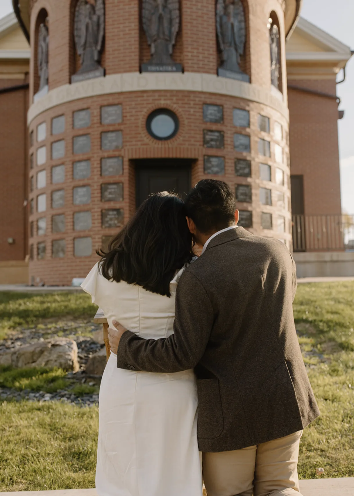 The couple embracing in front of a brick chapel, seen from behind