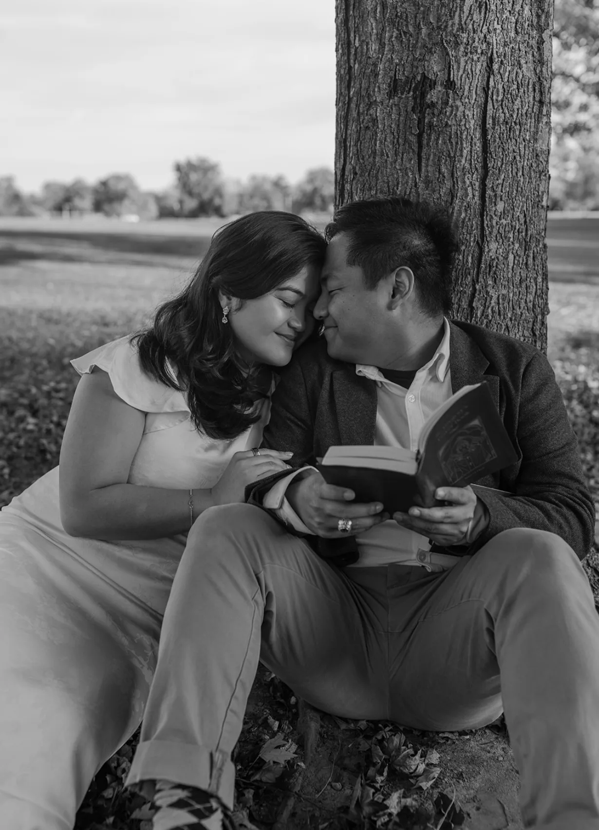 Black-and-white of the couple sitting under a tree, foreheads together, a book in his hand
