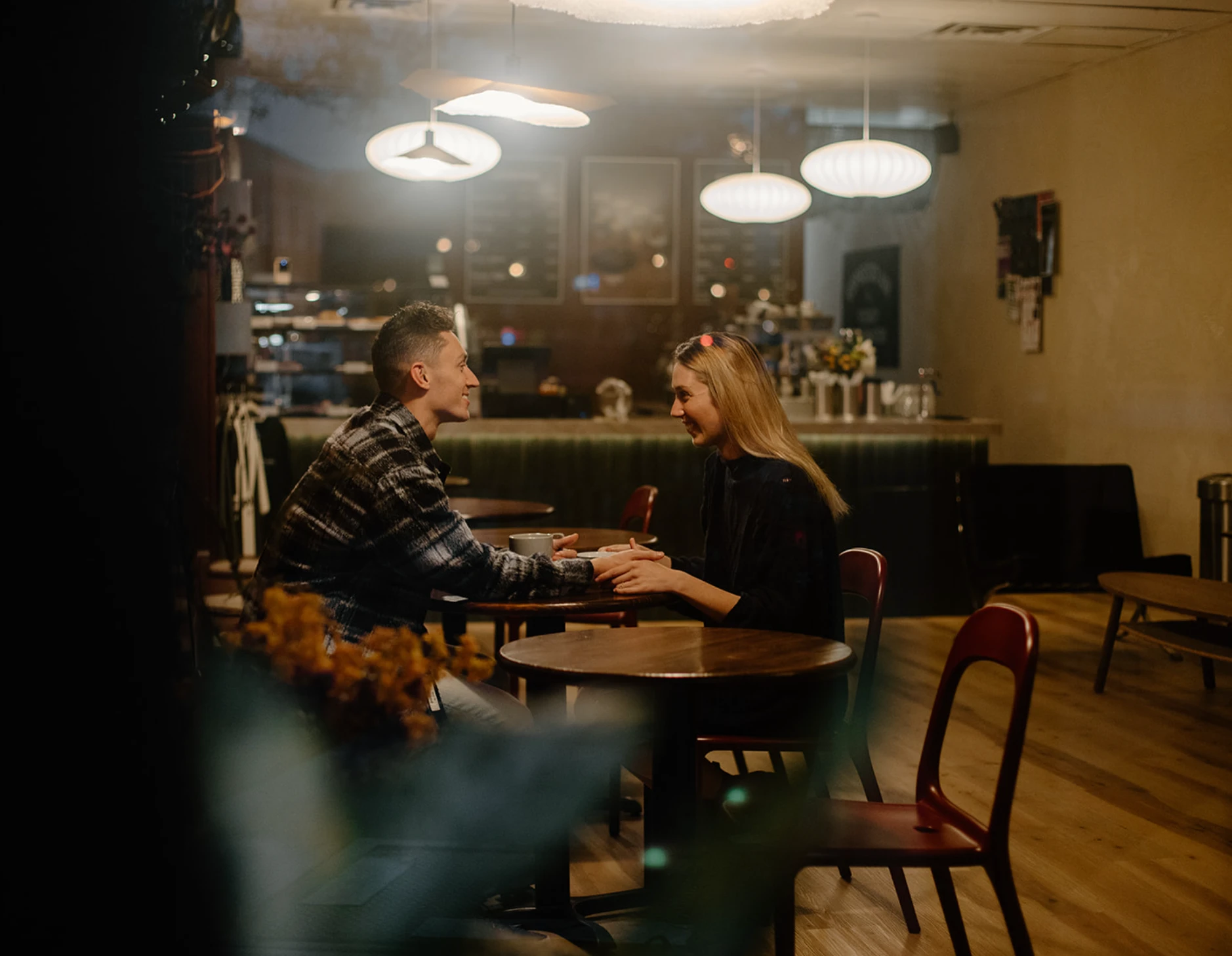 The couple holding hands at a café table, seen through the window at night