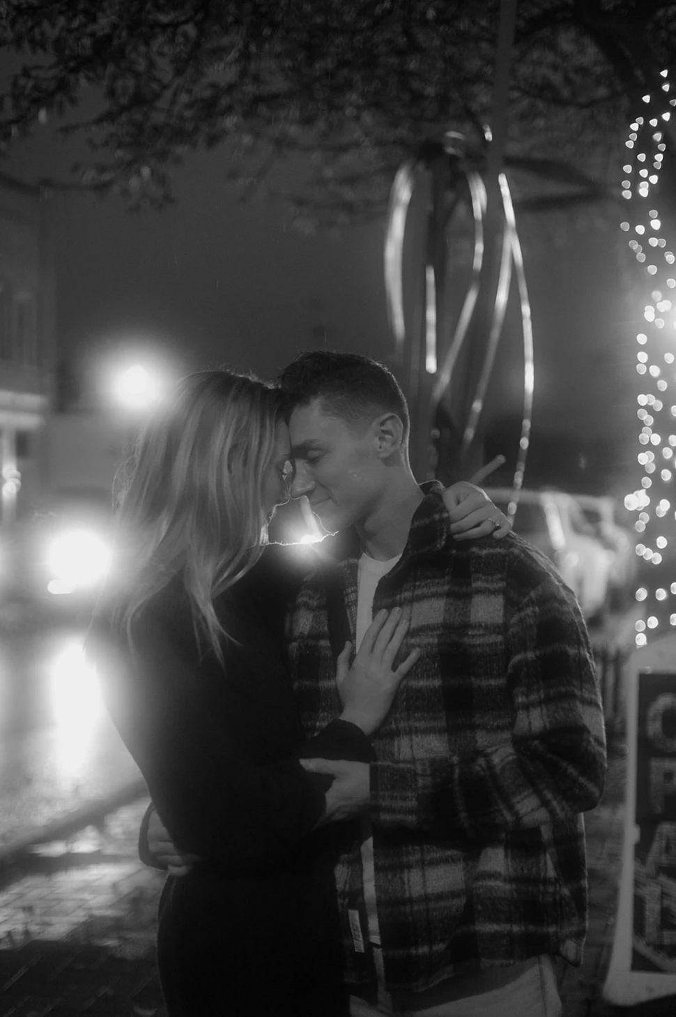 Black-and-white of the couple forehead to forehead under string lights on a rainy street