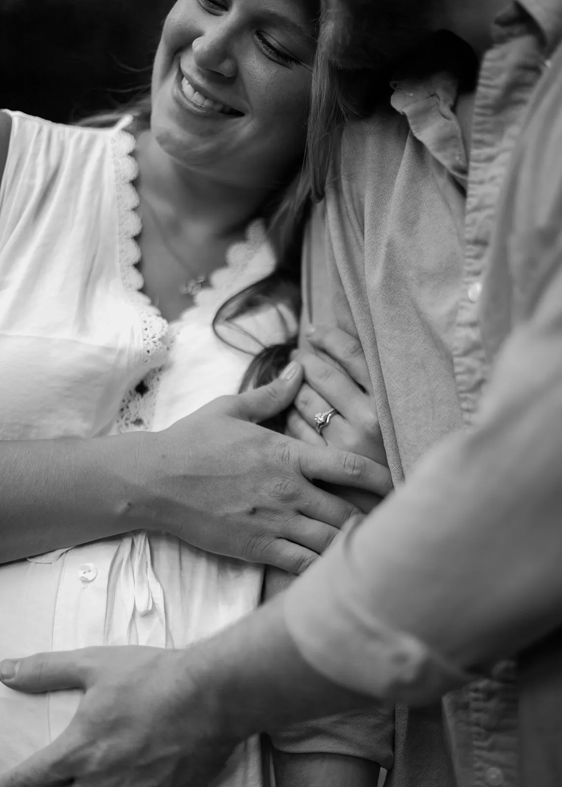 Black-and-white close-up of her smiling, resting on his shoulder with their hands on her belly