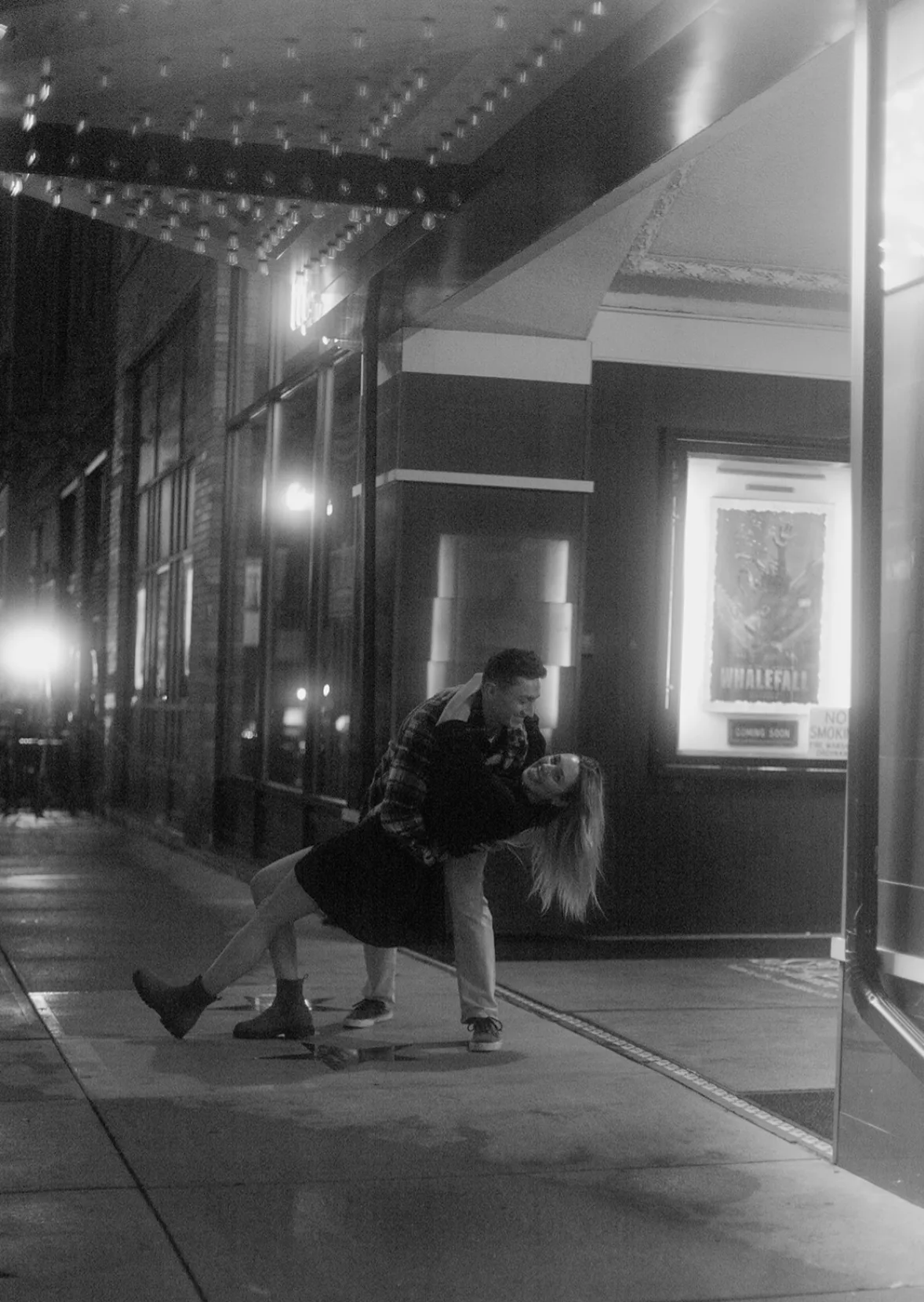 A couple dancing under a lit theater marquee at night, he dips her as she laughs