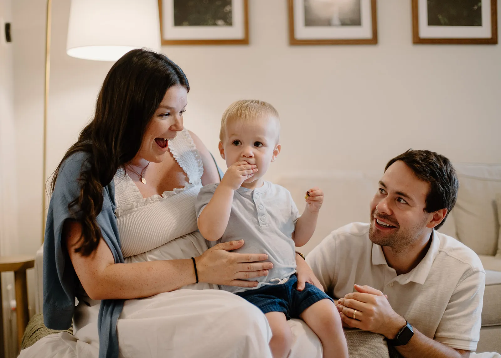 Mom laughing as their toddler snacks on her lap, dad smiling from the couch